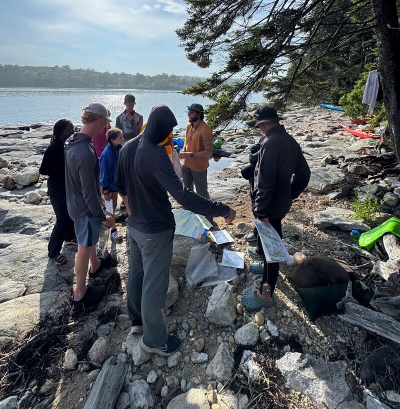 A group of people are gathered on a rocky shoreline near a body of water, possibly a lake or ocean. They appear to be looking at maps or documents spread out on the ground. The group consists of men and women of varying ages. The weather seems sunny, and the scene suggests an outdoor activity, perhaps related to navigation or exploration.
